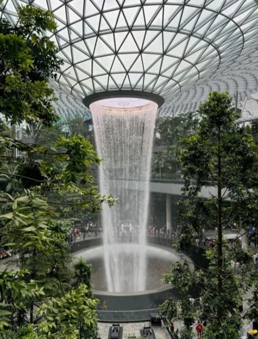 A stunning indoor waterfall surrounded by lush greenery and visitors, within a modern architectural space featuring a glass dome.